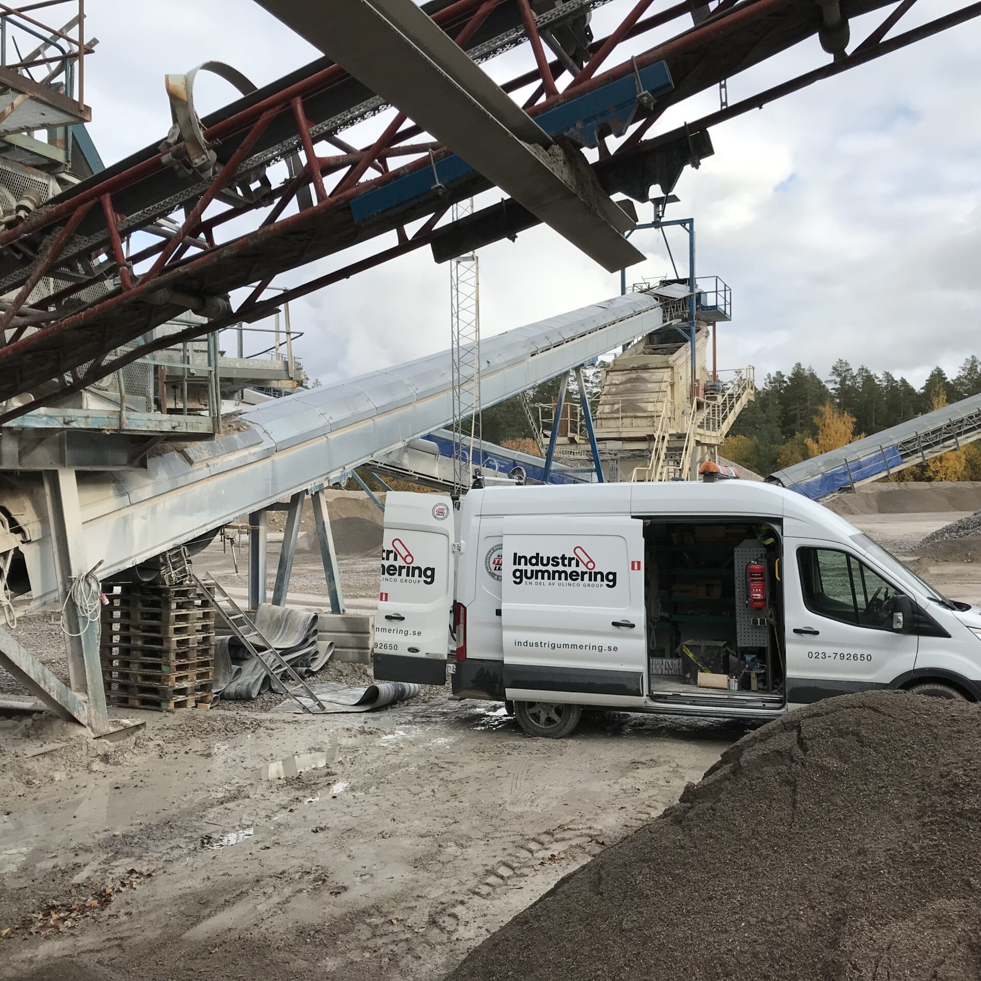 Servicebil från Industrigummering parkerad vid industriområde med transportband och grusmaterial i bakgrunden.